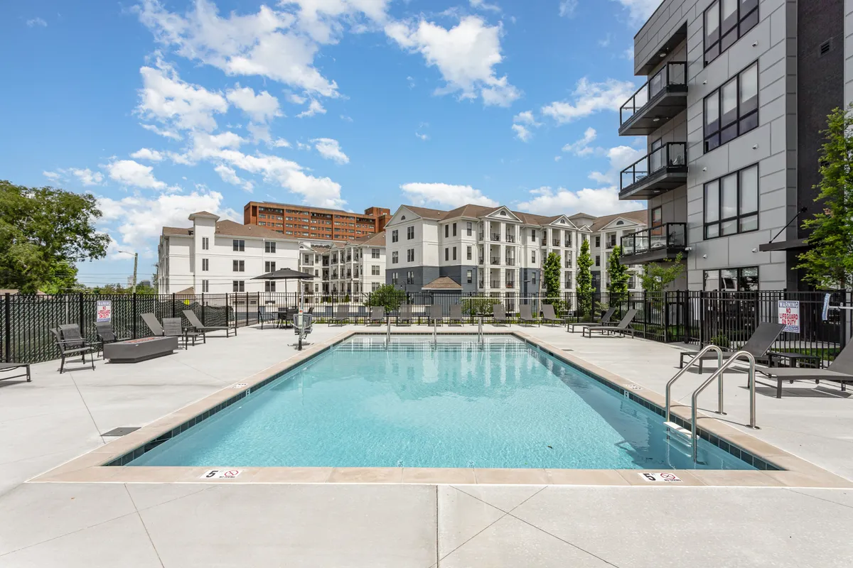 A sparkling swimming pool with an expansive lounge area framed by modern apartment buildings. The space is designed for sunbathing, relaxing, and gathering with friends and family, evoking a sense of leisure and community events.
