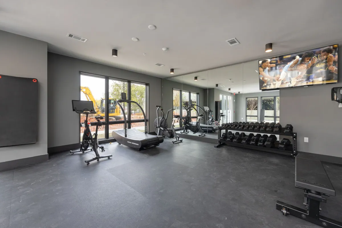 A modern fitness center featuring a treadmill, weight rack, exercise bike, large windows letting in natural light, and a television. The space is clean, bright, and designed for various types of workouts, fostering both individual fitness and a sense of community.