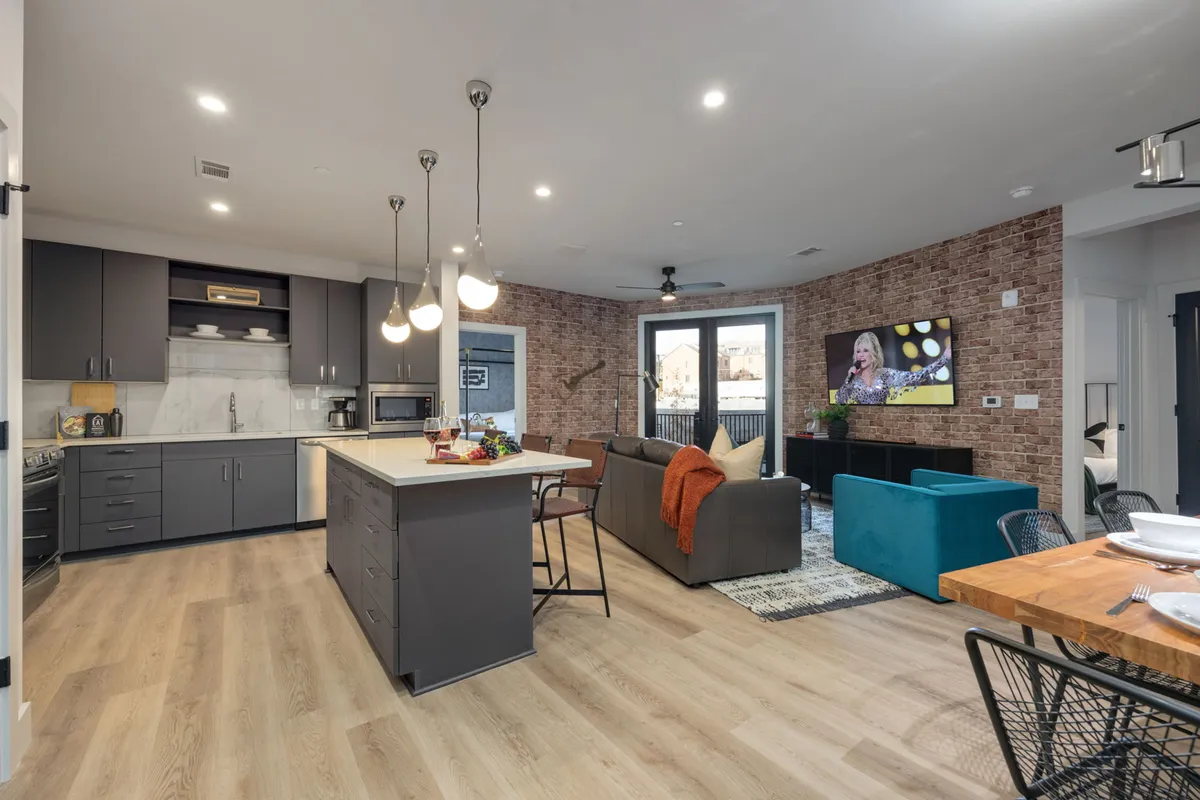 This image captures a spacious open-concept interior where the kitchen, dining area, and living room seamlessly flow together. Modern furnishings, a kitchen island, a plush sofa, and a large TV are visible, all flooded with natural light from expansive windows.