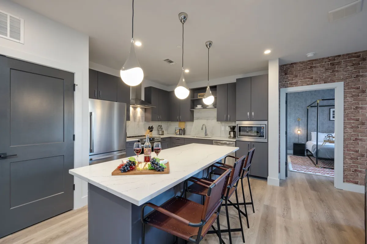 A light-filled, modern kitchen featuring a spacious island countertop adorned with wine and grapes, set beneath chic pendant lighting. The backdrop includes stainless steel appliances and a striking brick accent wall, creating a perfect space for cooking, entertaining, and gathering with friends or family.