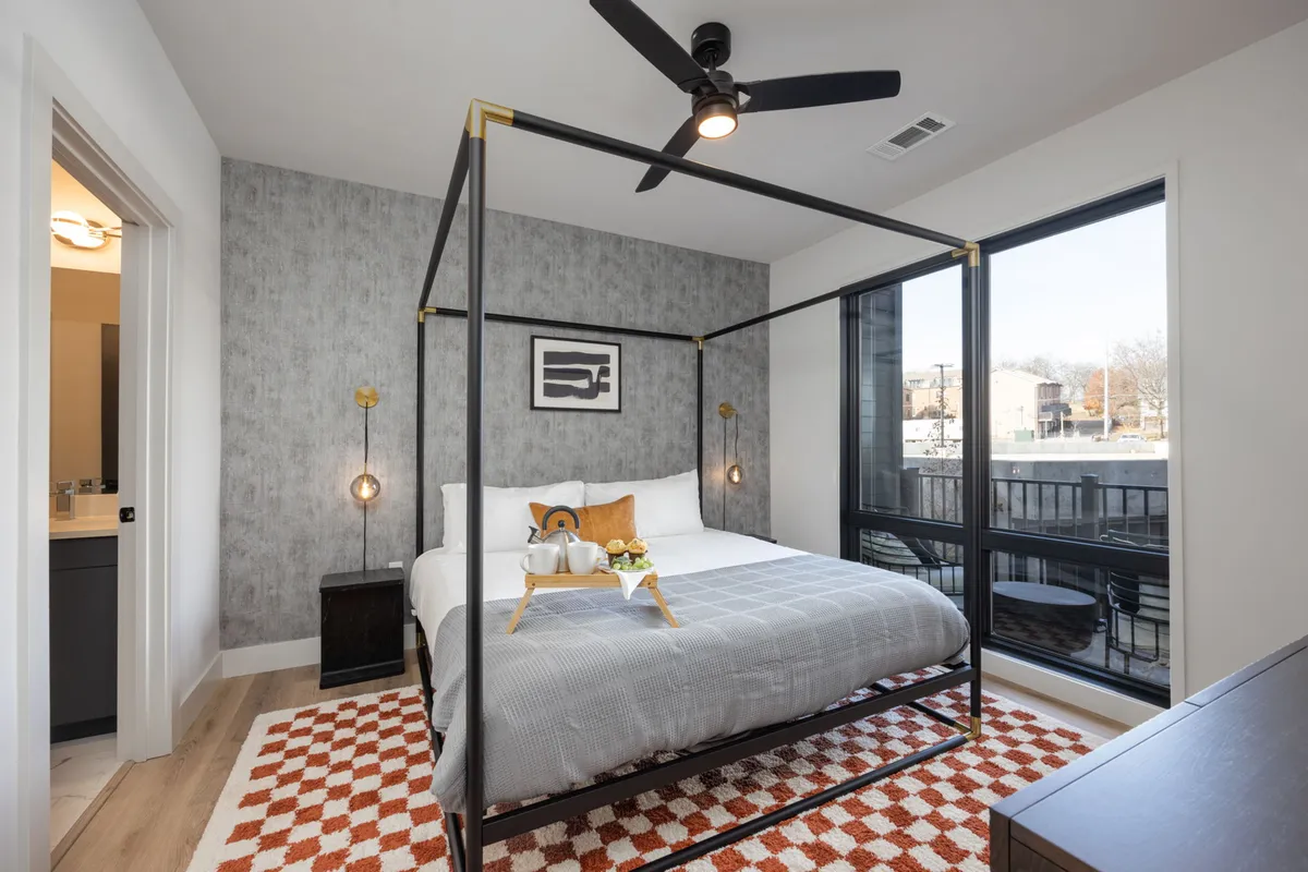 A stylish primary bedroom with a four-poster king bed dressed in crisp linens and decorative pillows. Sunlight streams through a nearby window, illuminating the comfortable, modern setting. The room is spacious and offers a serene atmosphere, ideal for restful nights and relaxed mornings.
