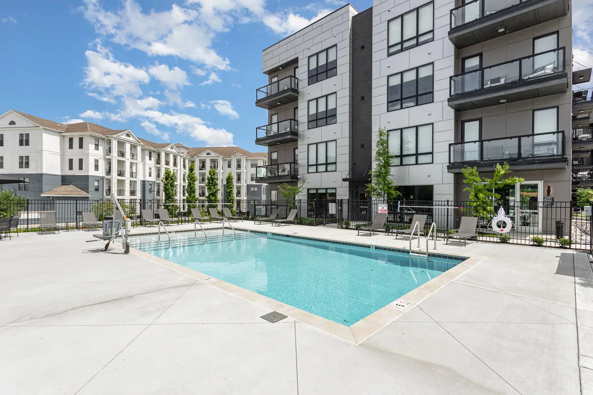 A year-round swimming pool surrounded by modern apartment buildings, lush green landscaping, and sun loungers. The setting is inviting for swimming, sunbathing, or socializing, with an atmosphere that feels both luxurious and communal.
